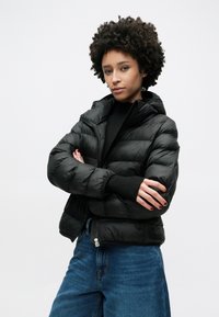 Μαύρο μπουφάν τύπου puffer με φερμουάρ μπροστά, ελαστικές μανσέτες και κουκούλα. Διαθέτει υφή με ραμμένα σχέδια και κοντό σχεδιασμό, συνδυασμένο με μπλε τζιν.