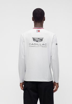 Homme de dos portant une chemise blanche à manches longues avec le logo de l'équipe Cadillac Formula 1 et un pantalon noir sur fond uni.