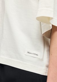 Λευκό πουκάμισο με κοντά μανίκια και μια μικρή ραμμένη ετικέτα με την ένδειξη «Marc O’Polo» κοντά στο τελείωμα, φορεμένο με μαύρο παντελόνι.