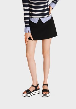 Donna che indossa una minigonna nera, un top a maniche lunghe a righe blu navy sovrapposto a una camicia a quadri blu e sandali platform neri con suola bianca.