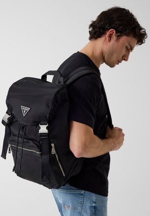 Jonge man draagt een zwart t-shirt en blauwe spijkerbroek met een zwarte Guess-rugzak met voorvakken met rits en gespsluitingen.