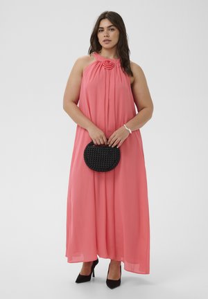 Kvinde iført en koralfarvet maxi kjole med halterneck og blomsterdetaljer, der holder en lille sort flettet håndtaske og har sorte hæle på.