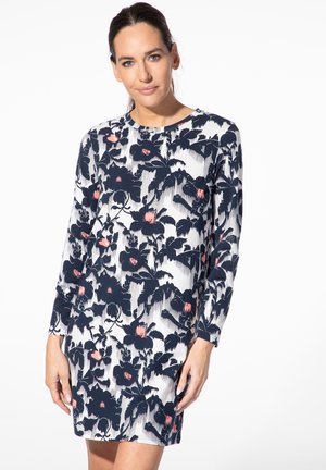 Frau trägt ein langärmliges weißes Kleid mit einem schwarzen und pinken Blumenmuster und steht vor einem einfarbigen weißen Hintergrund.