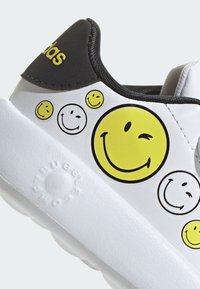 White sneaker with black accents and yellow smiley face designs. Features a textured rubber sole and an Adidas logo on the heel.
