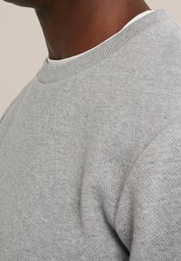 Svetle šedá mikina vyrobená z texturované látky, se zaobleným výstřihem a žebrovanými manžetami. Detailní záběr zdůrazňuje šití a detaily materiálu.