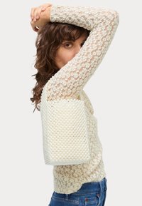 Borsa a tracolla con perline in bianco, dalla forma strutturata. Presenta una superficie testurizzata simile a perle e una base solida. Indossata sotto il braccio con un top di pizzo.