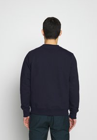 Felpa blu navy con maniche lunghe e polsini a coste, caratterizzata da un colletto a girocollo. Indossata sopra pantaloni a quadri.