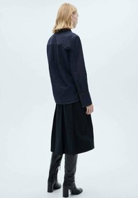 Chemise en denim bleu foncé, avec des manches longues et des poignets boutonnés, associée à une longue jupe noire et des bottes noires à talons montantes.