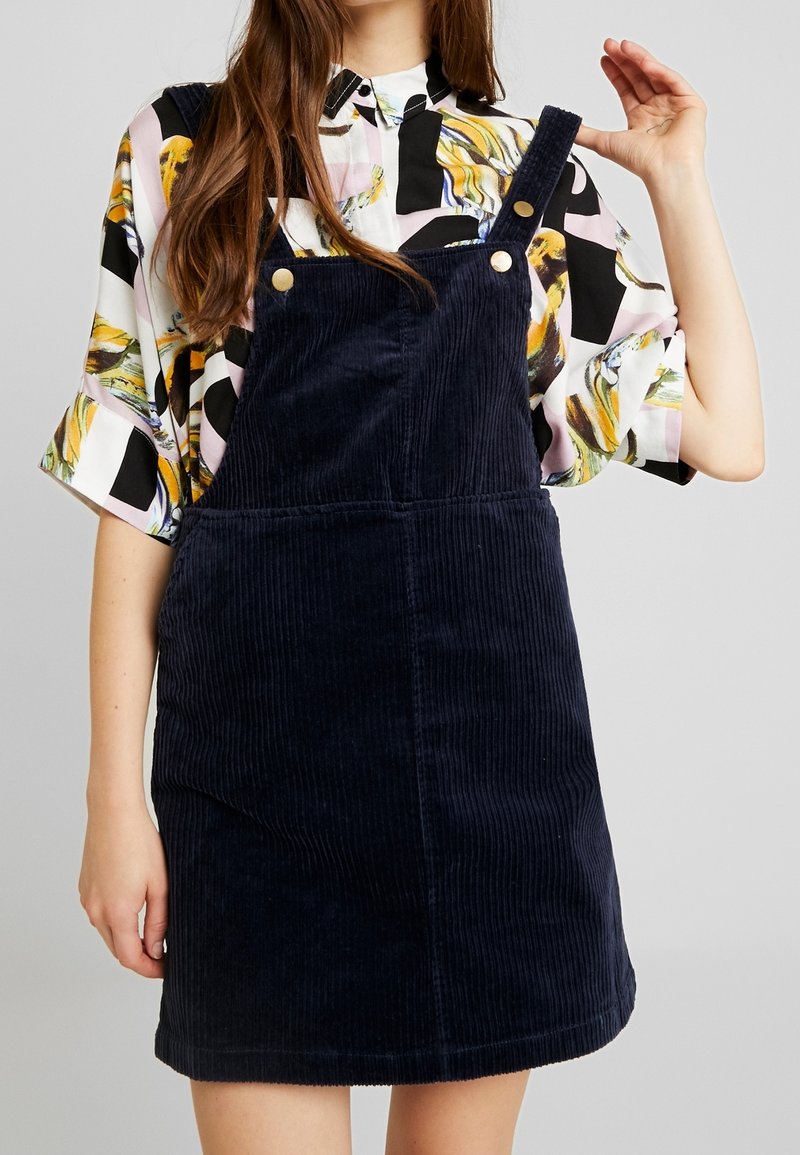 Robe salopette en velours côtelé bleu marine avec bretelles réglables et accents de boutons dorés, assortie à un blouse à manches courtes à motif.