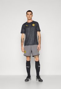 Sort mønstrede træningsskjorte med gul logo, grå shorts og sorte sokker. Har Puma-branding og et slående design.