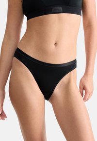 Vrouw in zwarte naadloze bikini-achtige onderbroek en bijpassende zwarte sportbeha, weergegeven van het midden van de romp tot boven de dijen.