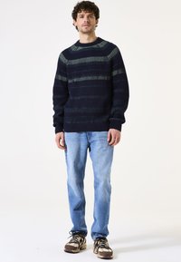 Suéter de punto azul marino con rayas horizontales verdes, textura acanalada, escote redondo, combinado con vaqueros azul claro y zapatillas beige.