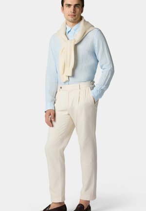 Hellblau gestreifte Bluse, cremefarbener Pullover um den Hals gebunden, cremefarbene Hose und dunkle Schuhe. Maßgeschneiderte Passform mit Falten und seitlichen Taschen.