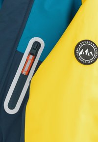 Giacca outdoor colorata con sezioni gialle, blu e blu navy. Presenta una tasca con cerniera con dettagli arancioni e una toppa rotonda con logo nero.