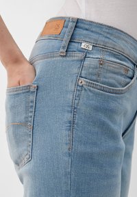 Jeans de mezclilla azul claro con corte slim, costuras de cobre, bolsillos laterales y un parche de logo de cuero marrón en la cinturilla trasera.