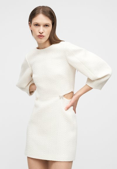 Γυναίκα που φοράει λευκό υφασμάτινο μίνι φόρεμα με φουσκωτά μανίκια και πλαϊνές περικοπές, στέκεται με τα χέρια στους γοφούς μπροστά σε μια απλή φόντο.