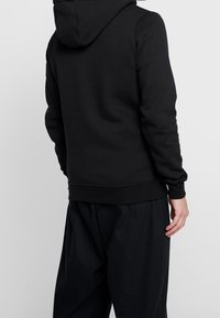 Muž v černé mikině s kapucí a volných černých kalhotách, stojící zády k fotoaparátu na jednoduchém šedém pozadí.