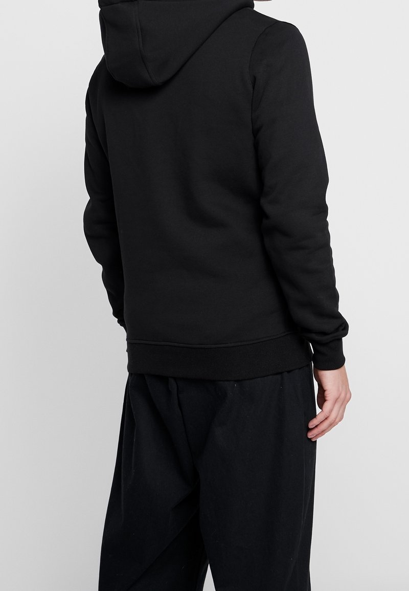 Muž v černé mikině s kapucí a volných černých kalhotách, stojící zády k fotoaparátu na jednoduchém šedém pozadí.