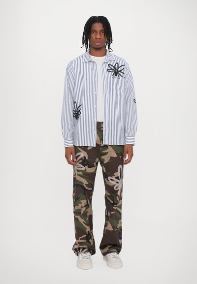 Stribet skjorte med sort blomsterbroderi, parret med camouflagedragtbukser med hvide blomsterdesigns, båret med hvide sneakers.