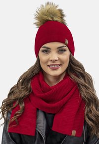 Bonnet en tricot rouge avec un pompon en fausse fourrure et une écharpe texturée assortie, tous deux présentant un motif subtil, accentué par une petite étiquette de marque.