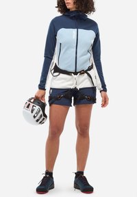 Person wearing a blue and white zip-up jacket, navy shorts with climbing harness, holding a white and black climbing helmet, standing on white background.