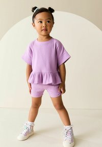 Lawendowy prążkowany dwuczęściowy strój z bluzką z baskinką i wzorzystymi szortami, wykonany z fakturowanej tkaniny oraz kolorowe trampki na sznurowadła.