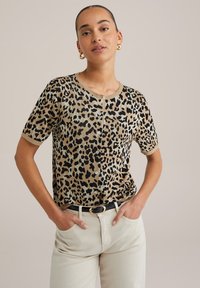 Haut en tricot à manches courtes au motif léopard beige et noir, avec un col rond et des détails de boutons. Accompagné d'un pantalon clair et d'une ceinture noire.
