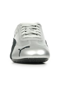 Zapatilla deportiva plateada con una superficie lisa y brillante, cordones blancos y detalles en negro, que cuenta con una suela de goma texturizada y un logotipo.