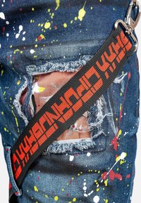 Jeans en denim avec un motif éclaboussé multicolore. Les zones usées montrent des bords effilochés. Une sangle noire avec du texte rouge est visible sur le côté.