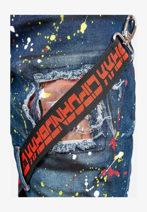 Jeans aus Denim mit mehrfarbigem Spritzmuster. Abgenutzte Stellen zeigen ausgefranste Kanten. Ein schwarzes Band mit rotem Text ist an der Seite sichtbar.