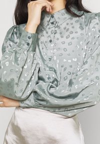 Silkegrøn bluse med krave, puffede ærmer og sølvfarvet leopardprintmønster; har matchende knapper langs fronten.