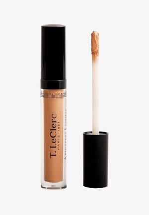 Flüssiger Concealer in einem durchsichtigen Röhrchen mit schwarzem Deckel. Applikator mit weicher Spitze, Produktfarbe ist ein warmes Beige. Branding ist auf dem Röhrchen sichtbar.