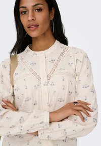 Femme aux longs cheveux bruns portant un chemisier crème à imprimé floral bleu avec des détails en dentelle, boucles d'oreilles créoles et une bague avec une pierre noire, les bras croisés.