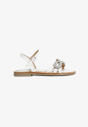 Sandali bianchi in pelle con suola piatta, cinturino alla caviglia regolabile e una decorazione in rilievo di colore argento a forma di nodo sulla parte anteriore.