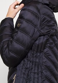 Chaqueta acolchada negra con paneles cosidos en horizontal, fabricada en un nylon brillante, con bolsillos laterales con cremallera y un logo decorativo en el hombro.