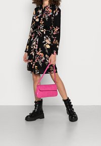 Kobieta w czarnej, kwiatowej sukience i czarnych butach na klamrę, trzymająca jaskraworóżową torebkę na tle gładkiej białej ściany.