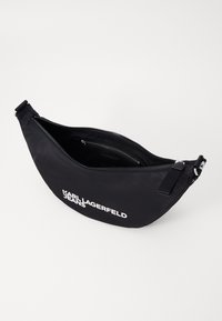 Borsa a tracolla nera in nylon con zip diagonale, caratterizzata da una grafica bianca con marchio. L'interno include una tasca più piccola per l'organizzazione.