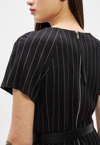 Moteris dėvi juodą suknelę su plonomis baltomis juostelėmis, trumpomis rankovėmis ir matoma sidabrine užtrauktuku nugaroje, prisegtą juodu diržu.