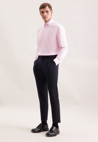 Uomo in piedi con le mani in tasca, che indossa una camicia da uomo rosa chiaro, pantaloni scuri su misura, calzini neri e scarpe eleganti in pelle nera lucida.