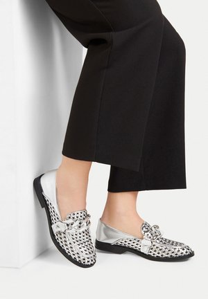 Silberne Slipper mit gewebtem schwarz-weißen Muster, ausgestattet mit einer dekorativen Schleife und einer flachen Sohle, vor schwarzen Hosen.