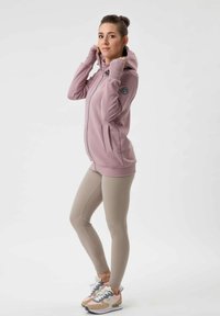Felpa leggera rosa con zip frontale, polsini a coste e patch con logo, abbinata a leggings beige e sneakers multicolore.