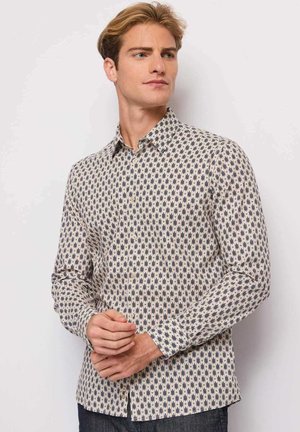 Giovane uomo con capelli biondi che indossa una camicia a maniche lunghe beige e blu navy con motivo a fantasia, in piedi davanti a uno sfondo bianco uniforme.