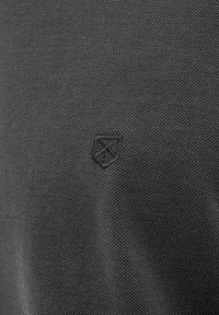 Detailný záber na tmavú tkaninu s textúrovaným povrchom a malým, embosovaným logom štítu. Dizajn je minimalistický a moderný.