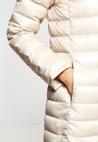 Pufová bunda s lesklým, světle béžovým povrchem, prošívanými sekcemi a otevřenou kapsou na boku, která má hladkou strukturu.