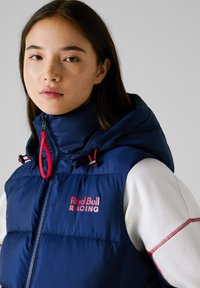 Μπλε ναυτικό γιλέκο με κουκούλα, που διαθέτει μπροστινό φερμουάρ, κόκκινες λεπτομέρειες και το λογότυπο "Red Bull RACING" στο στήθος. Απαλή υφή, ελαφριά σχεδίαση.