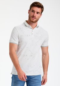 Polo bianco con un motivo testurizzato, colletto con due bottoni e dettagli neri, abbinato a jeans azzurri chiari.