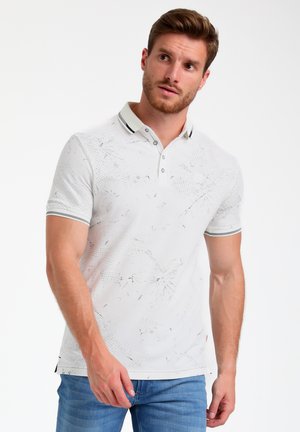 Polo bianco con un motivo testurizzato, colletto con due bottoni e dettagli neri, abbinato a jeans azzurri chiari.