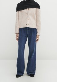 Beige gestrickte Strickjacke mit Rundhalsausschnitt und Knöpfen, drapiert mit einem schwarzen Schal. Kombiniert mit lockeren blauen Jeans.