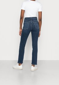 Pantalones vaqueros de mezclilla azul oscuro con cintura alta, corte ajustado, diseño de cinco bolsillos y costuras en contraste, combinados con zapatillas blancas.