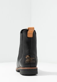 Botine negre din piele cu elastice pe laturi, un tab de tras din piele maro deschis și un patch cu logo maro, având o talpă din cauciuc texturat.